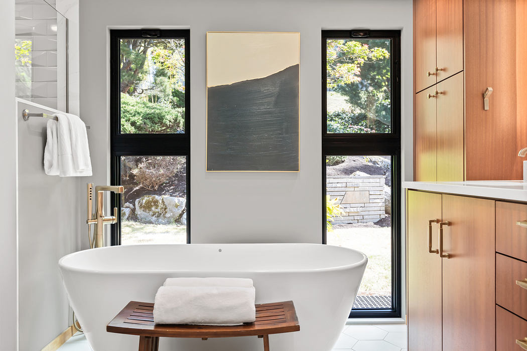 A modern bathroom with a freestanding tub, floor-to-ceiling windows, and wood cabinetry.