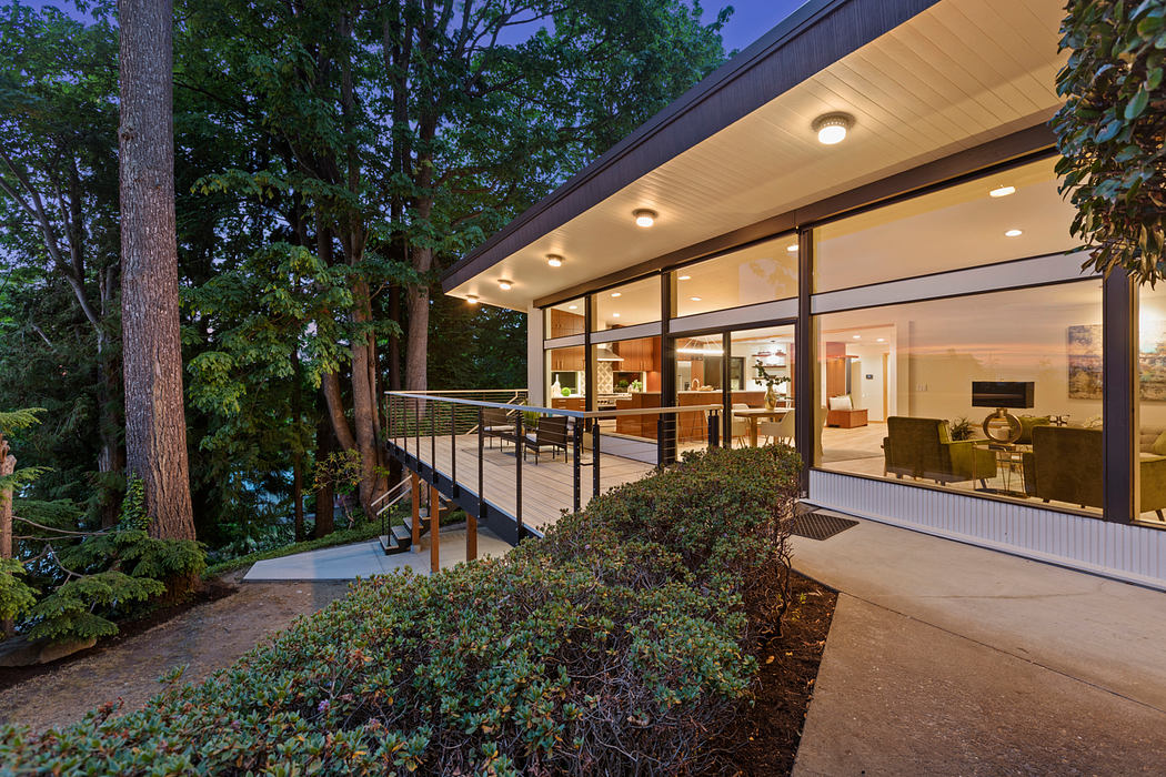 A modern, glass-walled home nestled in a lush, wooded setting, with a covered patio and deck.