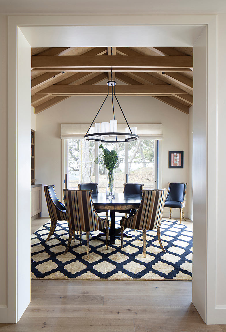A modern dining room with wooden beamed ceiling, chandelier, and patterned area rug.