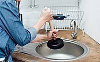 Partial view of woman in denim shirt using plunger in kitchen