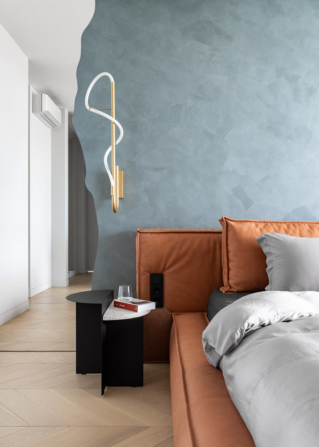 A modern bedroom with a gray concrete wall, a golden curved wall sconce, and a cozy brown leather sofa.