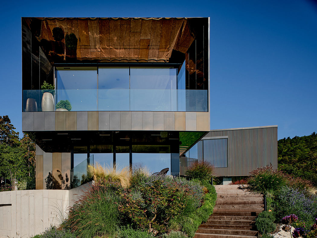 A modern, glass-walled structure with a wooden roof, surrounded by a lush, landscaped garden.