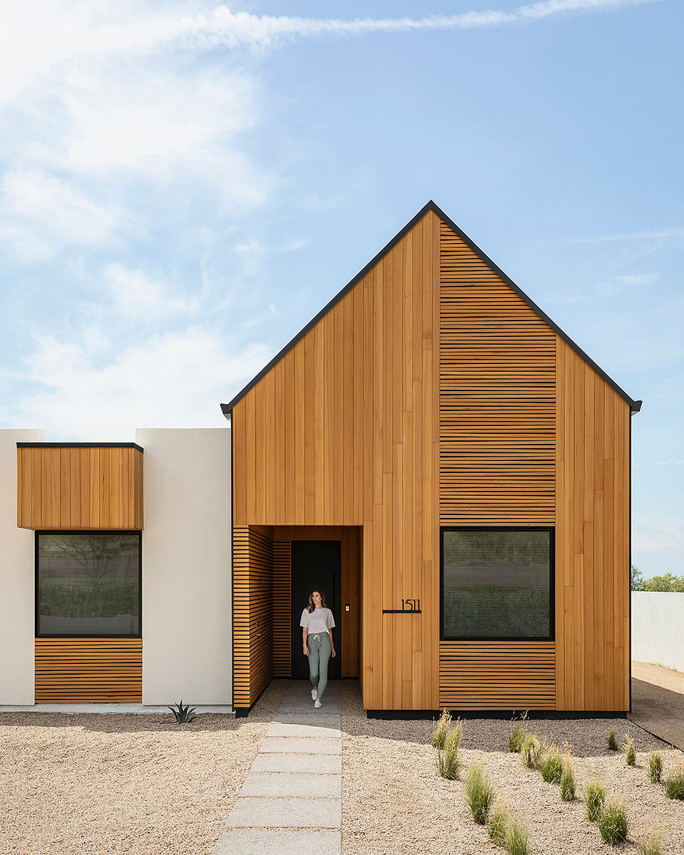 A wooden-clad modern home with a striking gabled roof and large windows, surrounded by a gravel path.