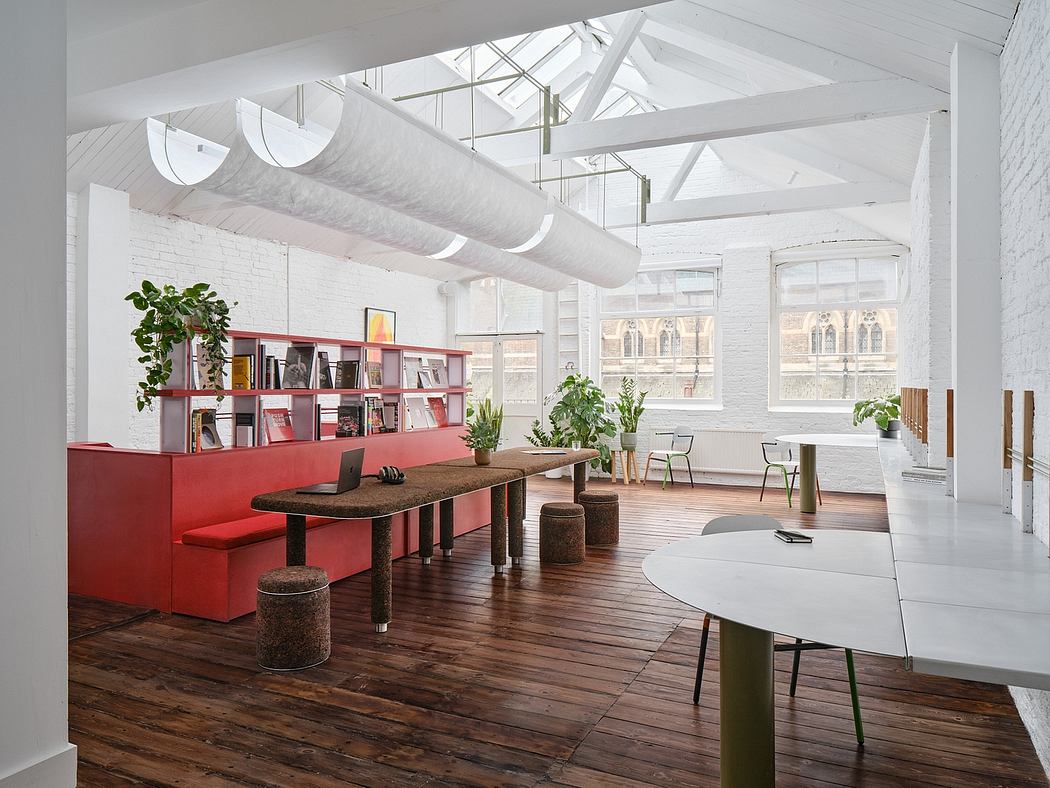 An open, industrial-style office space with red shelving, wooden furnishings, and large windows.