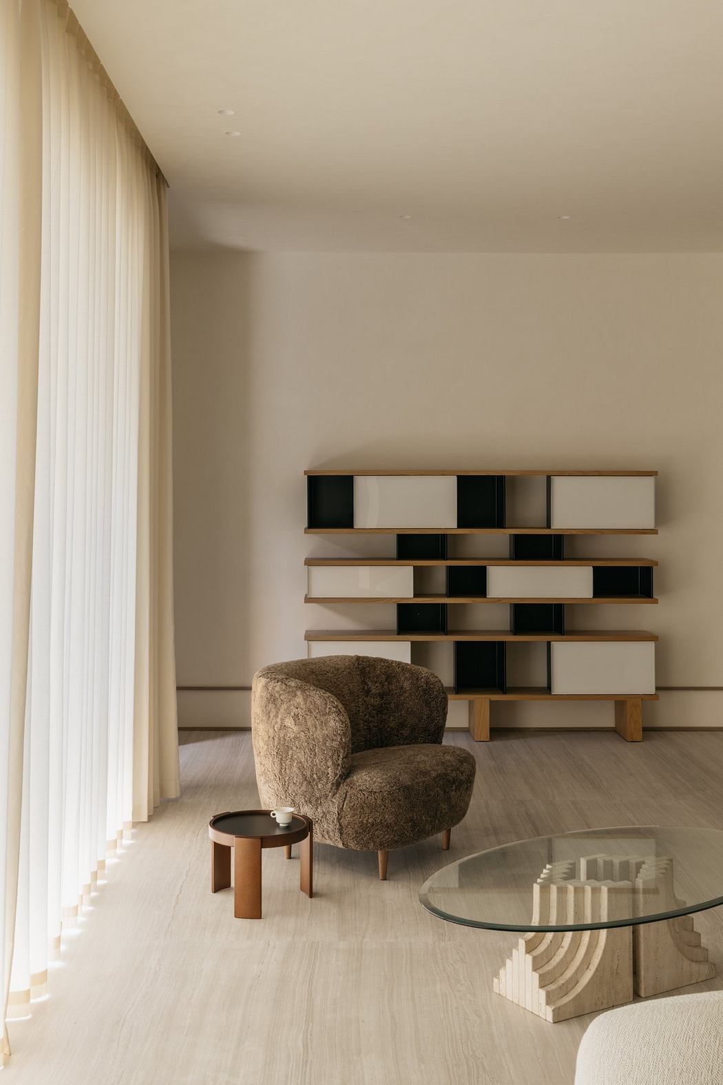 A modern living room with a minimalist wooden bookshelf, plush armchair, and glass-topped coffee table.