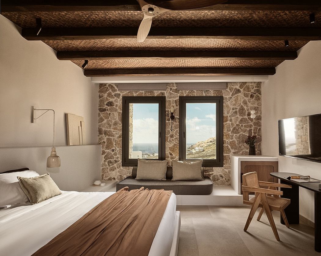 A cozy bedroom with a rustic stone wall, wooden beams, and panoramic windows overlooking a scenic landscape.