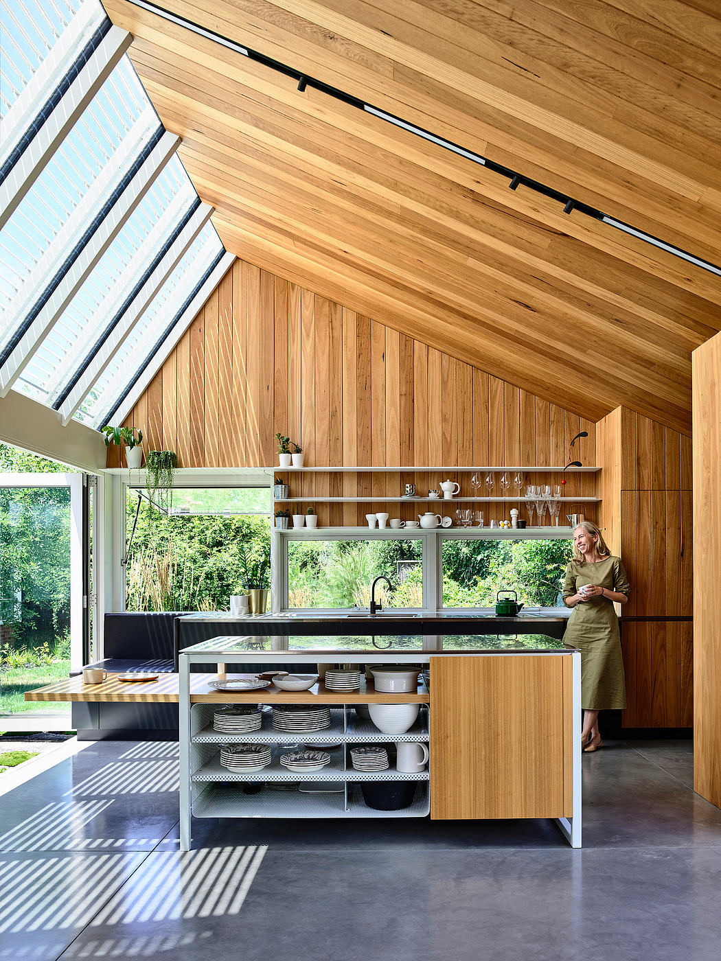 A modern kitchen with a wooden ceiling, shelving, and cabinetry, overlooking a lush outdoor view.