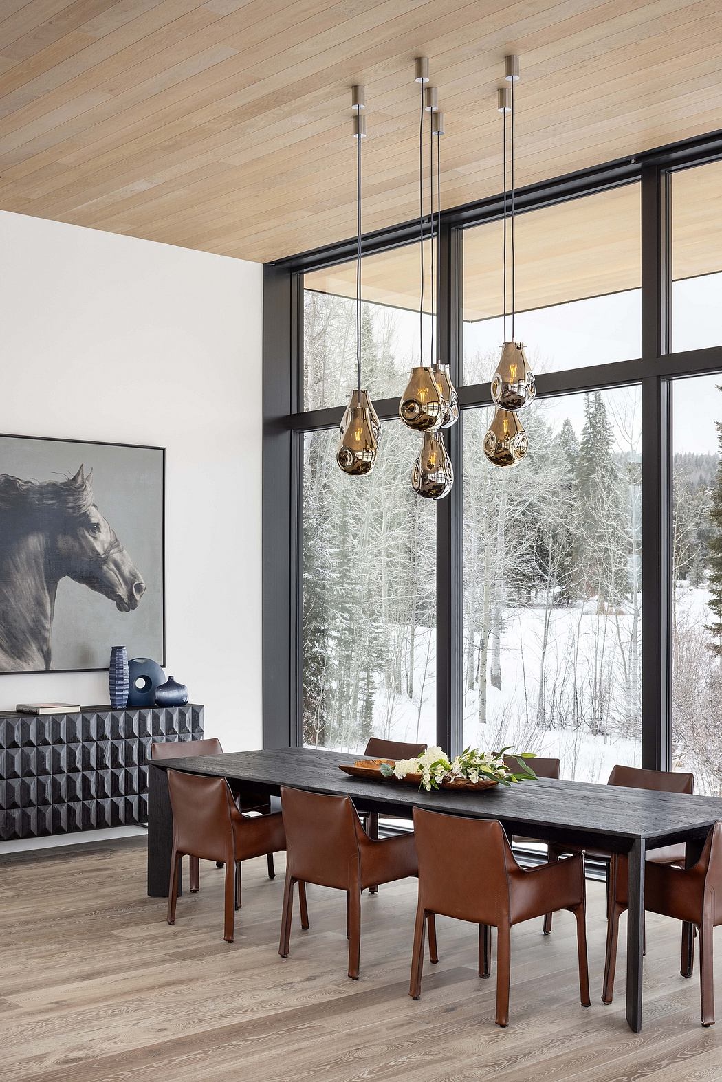 Cozy dining space with sleek wooden furniture, pendant lighting, and snowy forest view.