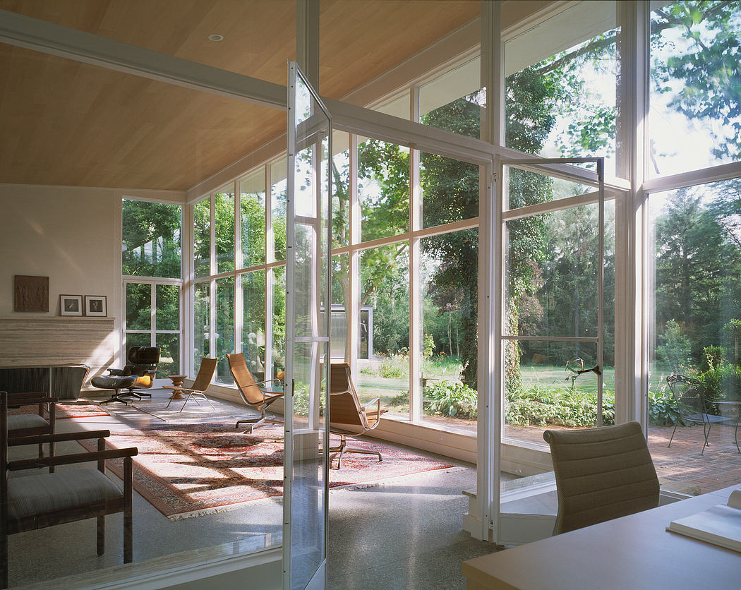 A spacious glass-walled room overlooking a lush, forested garden with mid-century furnishings.