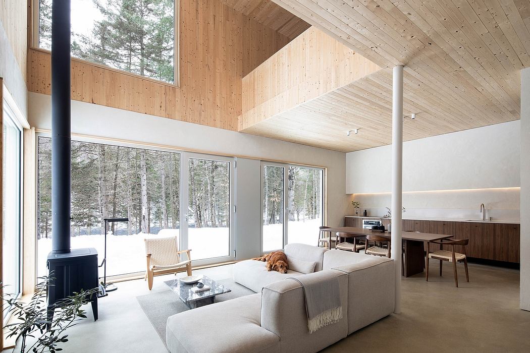 A cozy living area with large windows overlooking a snowy forest, featuring a wooden ceiling and minimalist decor.