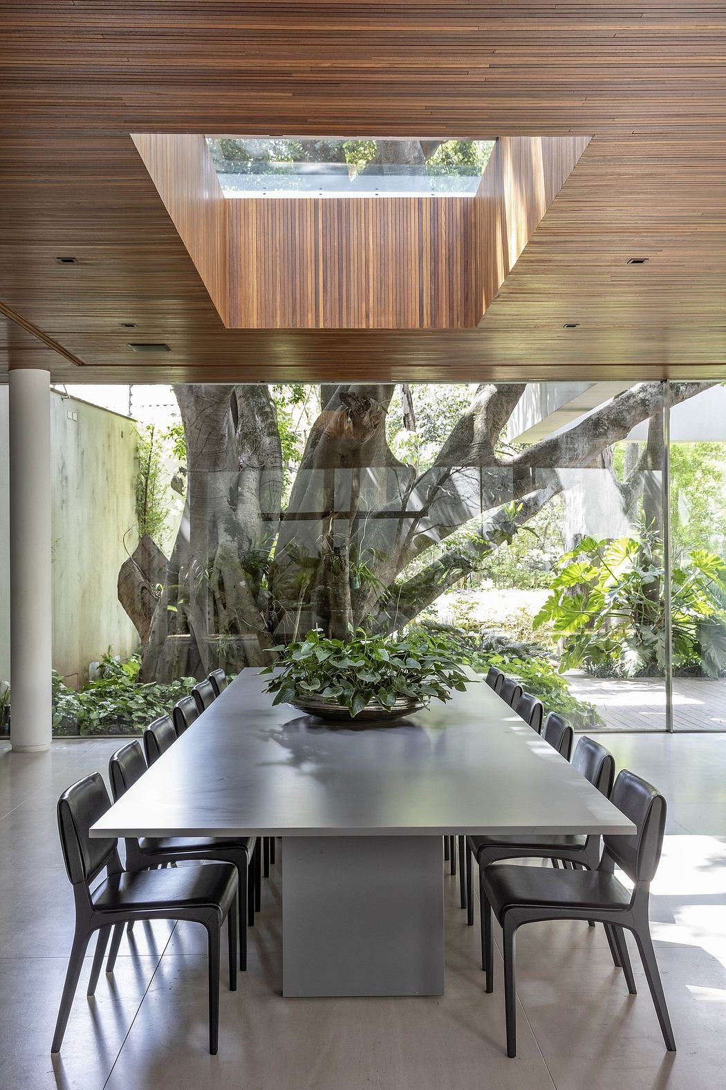 Wooden ceiling with skylight, glass walls, minimalist dining table and chairs, lush greenery.
