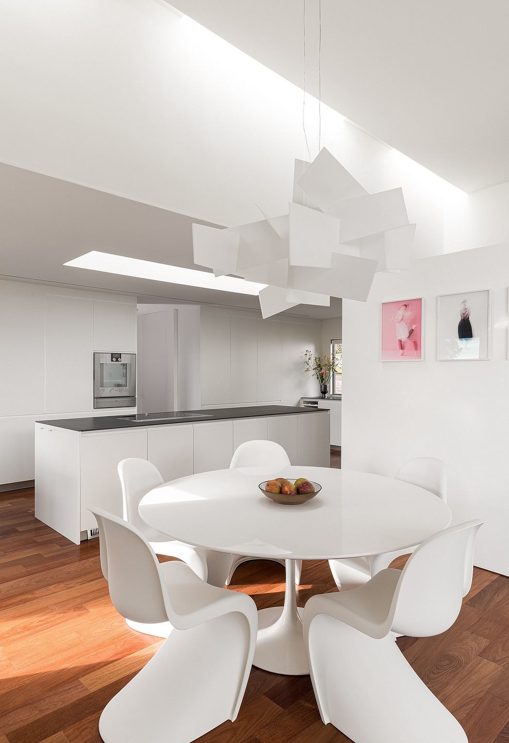A modern, minimalist kitchen and dining area with a sleek white table and chairs, accented by geometric light fixtures.