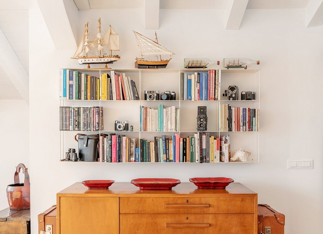 Shelves display books, cameras, and ship models against white walls and wooden dresser.