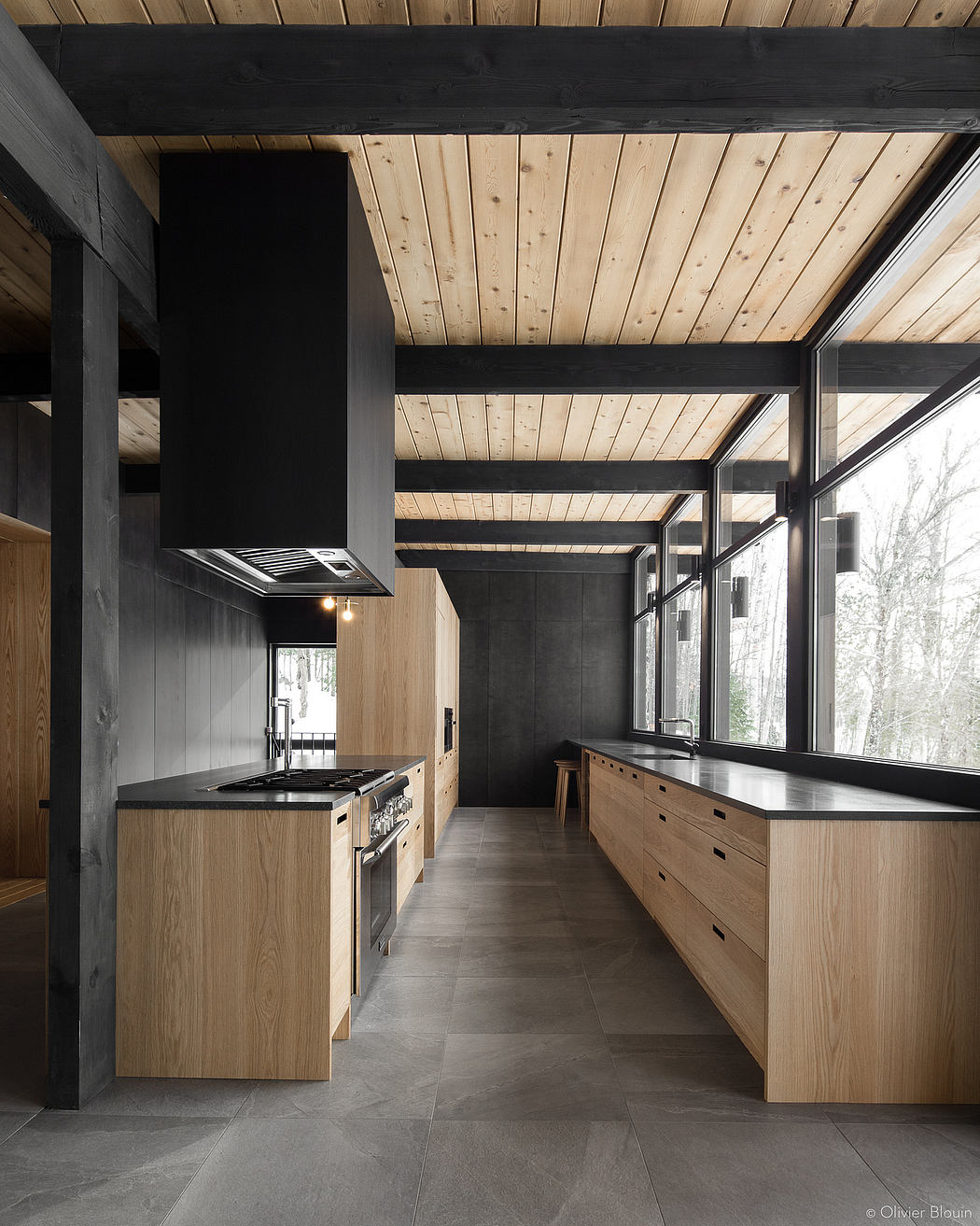 A modern rustic kitchen with dark wooden beams, light wood cabinets, and large windows.