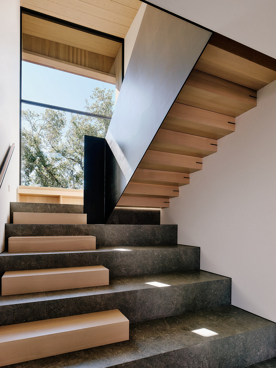 A modern staircase design with wood and stone materials, leading to a large window with natural light.