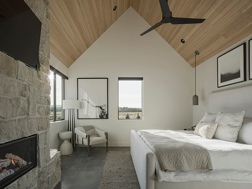 Cozy bedroom with vaulted wooden ceiling, stone fireplace, and panoramic window view.