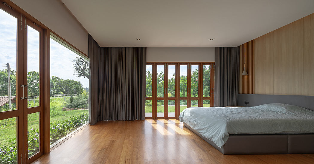 Modern bedroom with large windows overlooking lush greenery, wooden floors, dark curtains.
