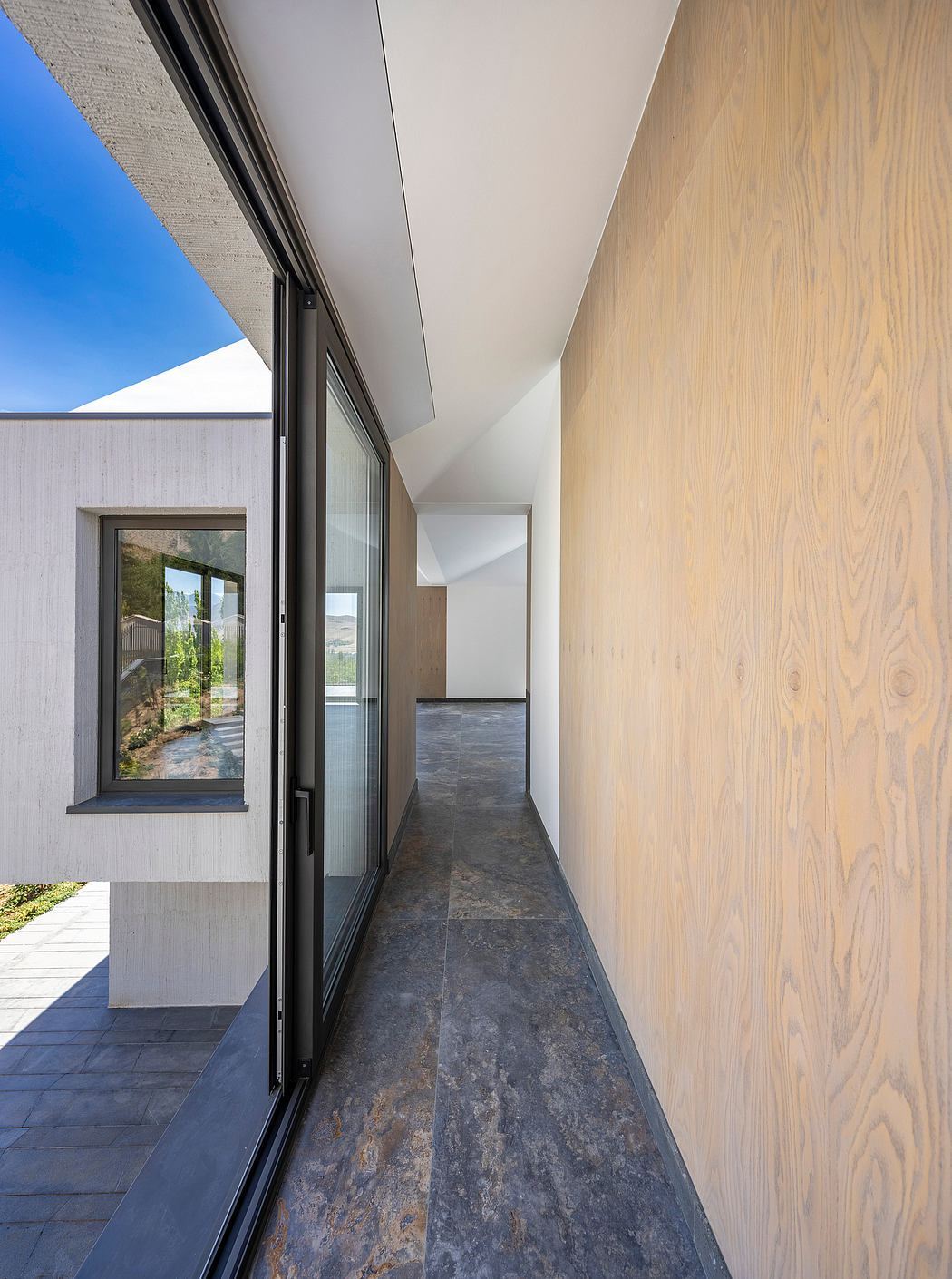 Spacious, minimalist hallway with wood-paneled walls, slate tiles, and floor-to-ceiling windows.