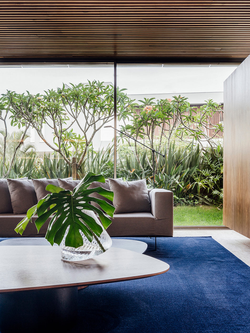A cozy living space with plush gray sofas, a wooden coffee table, and lush tropical foliage visible through the glass walls.