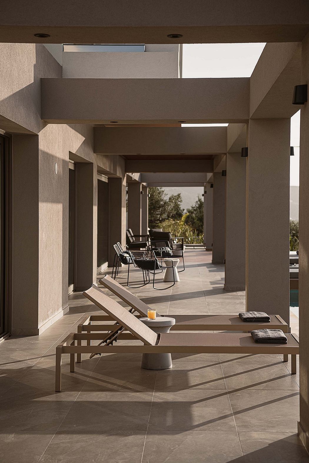 A minimalist outdoor patio with chic lounge chairs, a low table, and a tiled floor leading to a covered walkway.