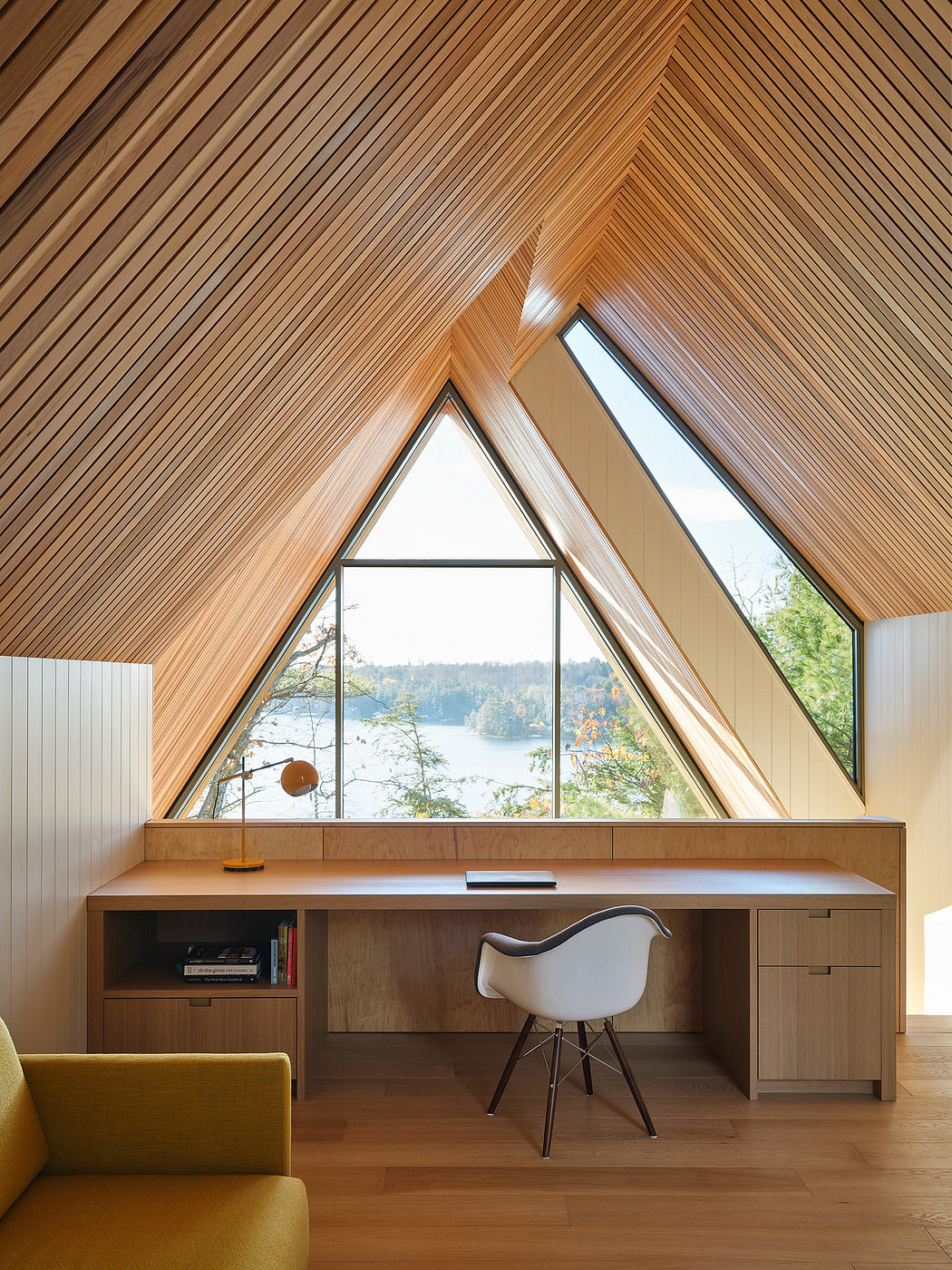A cozy home office with a triangular window framing a scenic lake view, surrounded by a warm wooden interior.