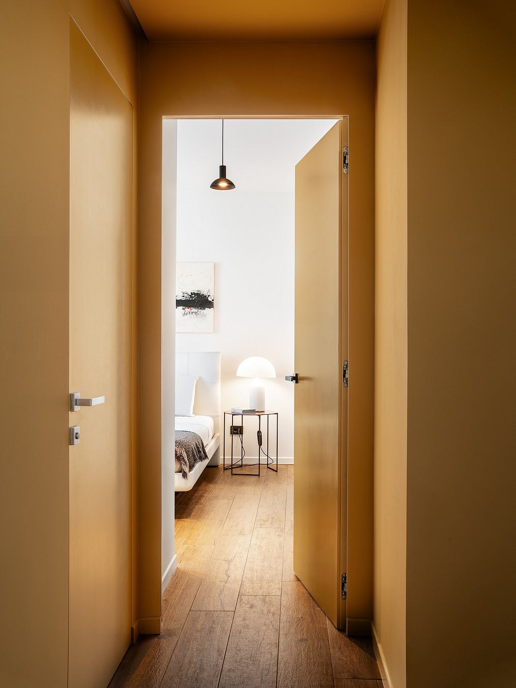 A warm, cozy bedroom glimpsed through an open doorway, with wooden floors and modern lighting.