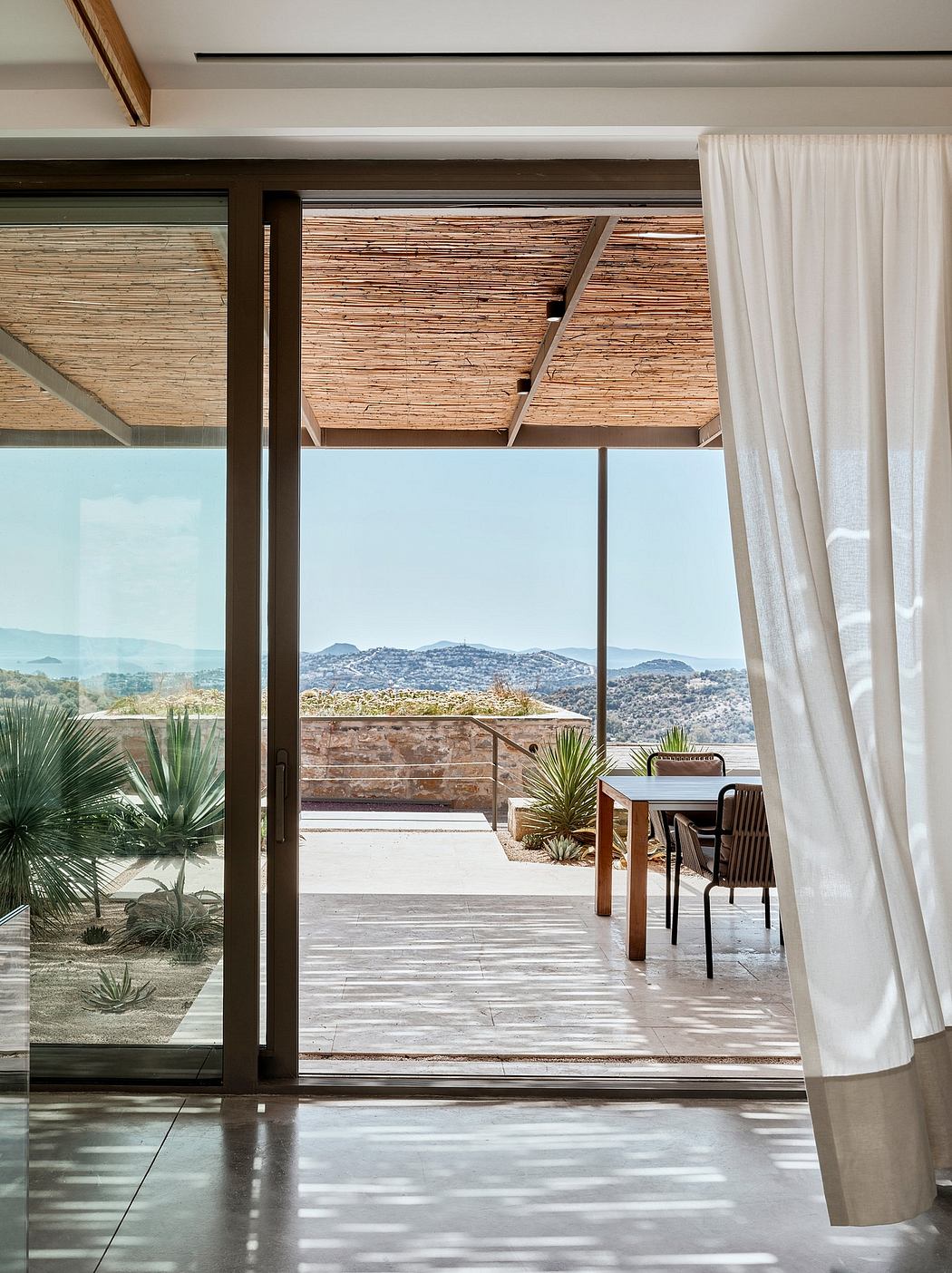 A modern indoor-outdoor space with a wooden slat ceiling, sliding glass doors, and desert landscaping.