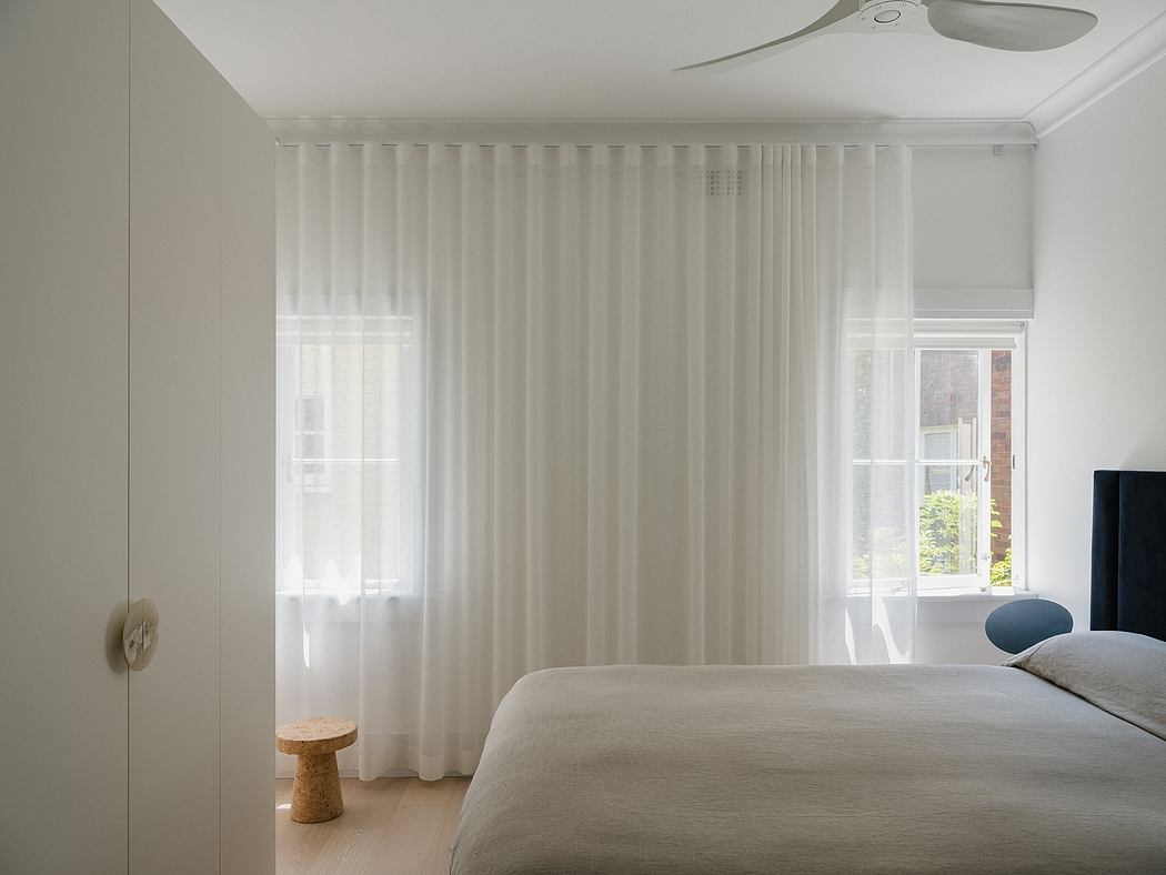 A bright, minimalist bedroom with floor-to-ceiling curtains, a ceiling fan, and a cork side table.
