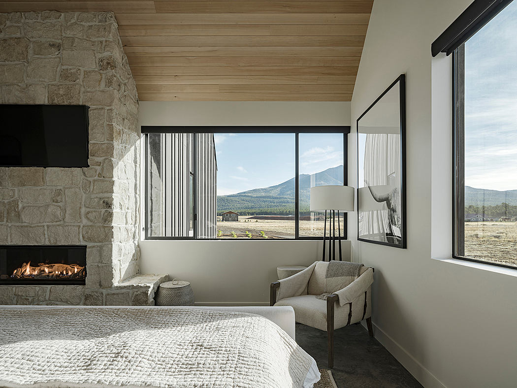 Rustic bedroom with stone wall, wooden beams, and panoramic mountain view through large windows.