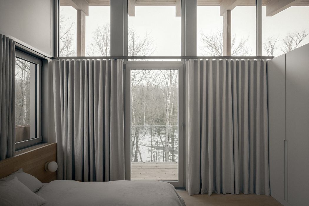 Minimalist bedroom with floor-to-ceiling windows, gray curtains, and a wooden deck overlooking a wintry landscape.