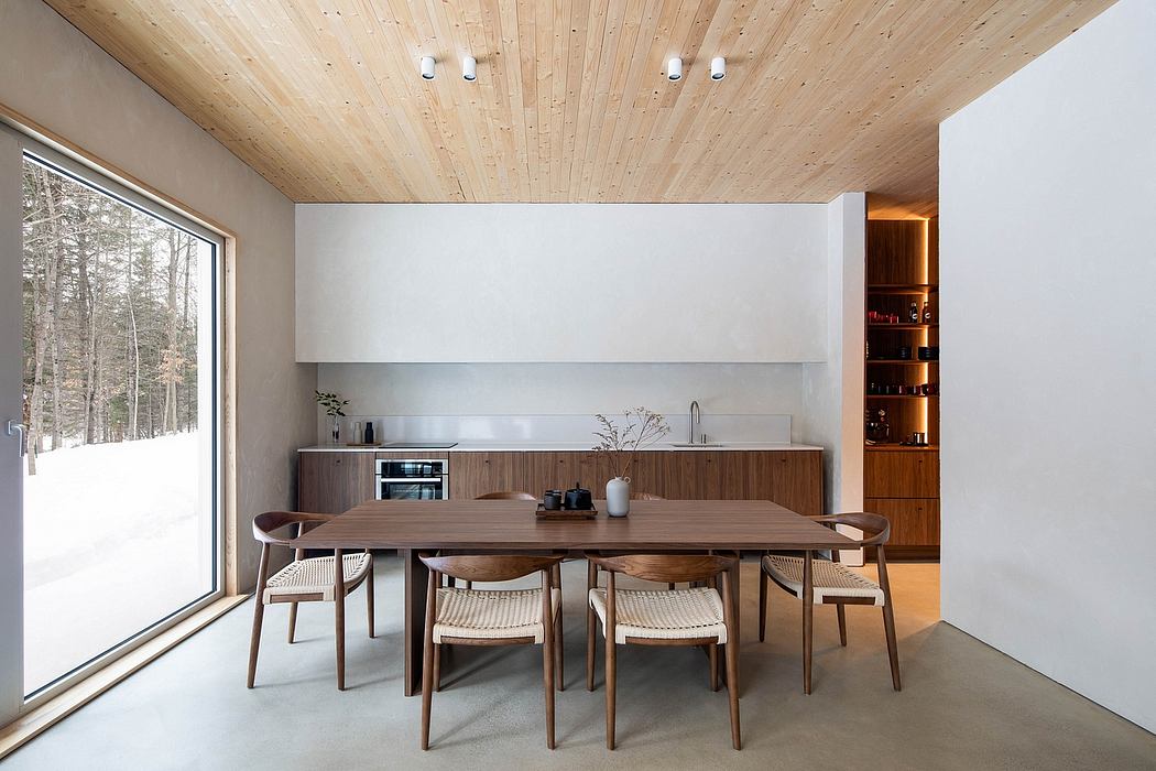 The image depicts a modern, wood-paneled interior with a large dining table, kitchen, and scenic window.