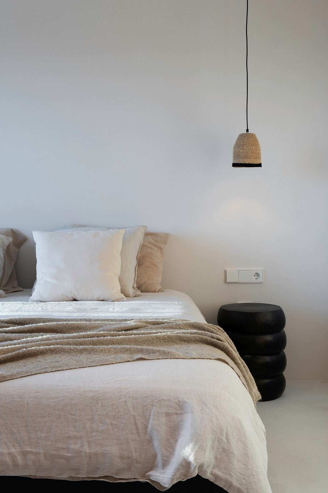 Minimalist bedroom with textured beige blanket, stacked black poufs, and pendant lamp.