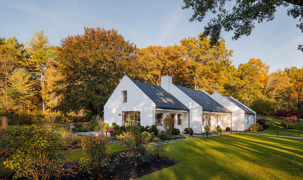 A charming white farmhouse with a pitched roof, surrounded by vibrant fall foliage and a lush lawn.