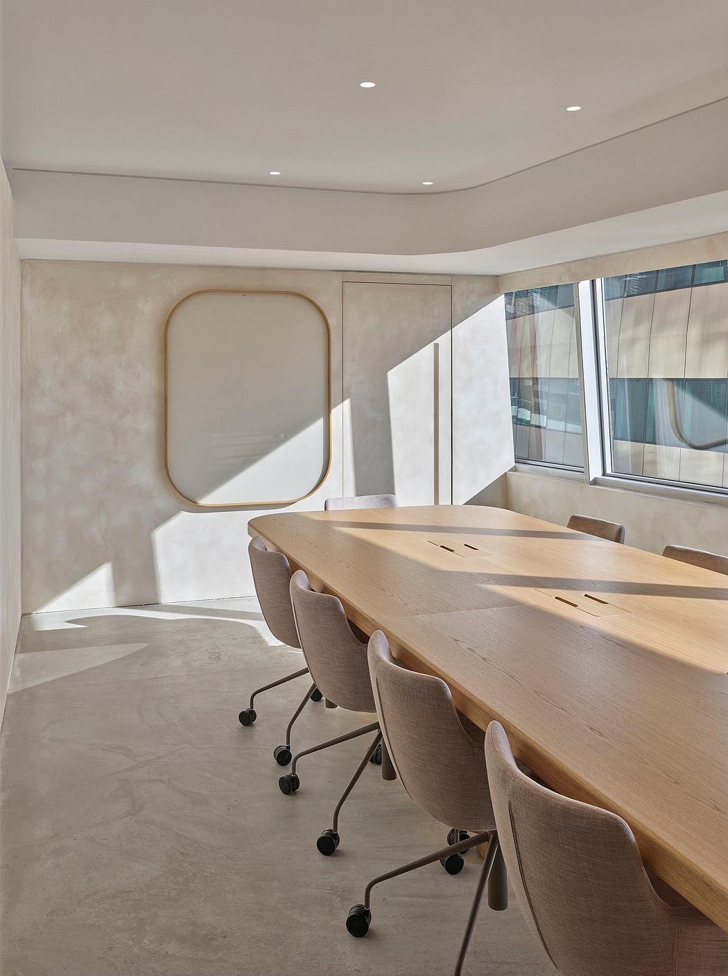 A modern, minimal conference room with a large wooden table, rolling chairs, and a large mirror on the wall.