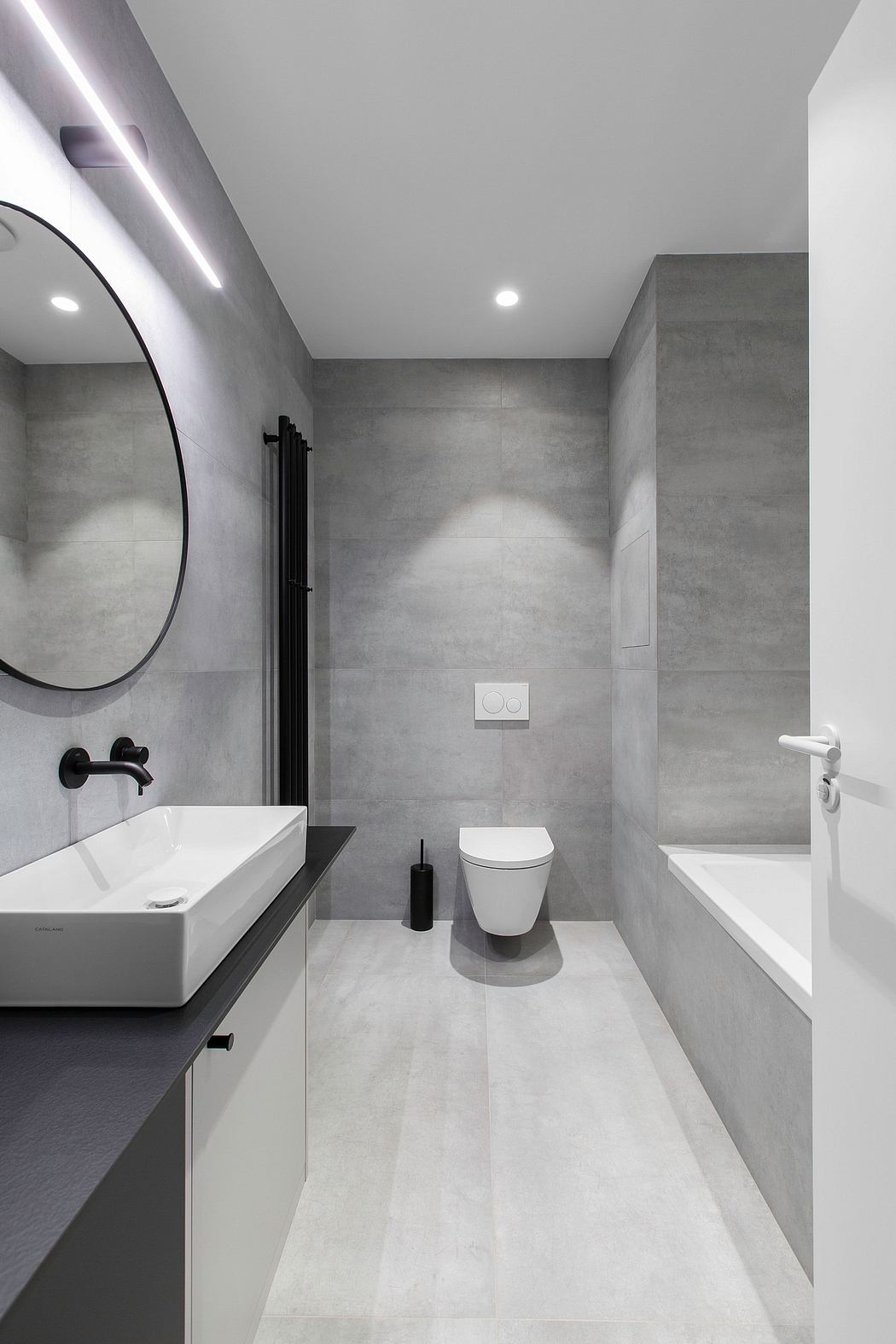 A modern, minimalist bathroom featuring sleek gray tile, a round mirror, and black fixtures.