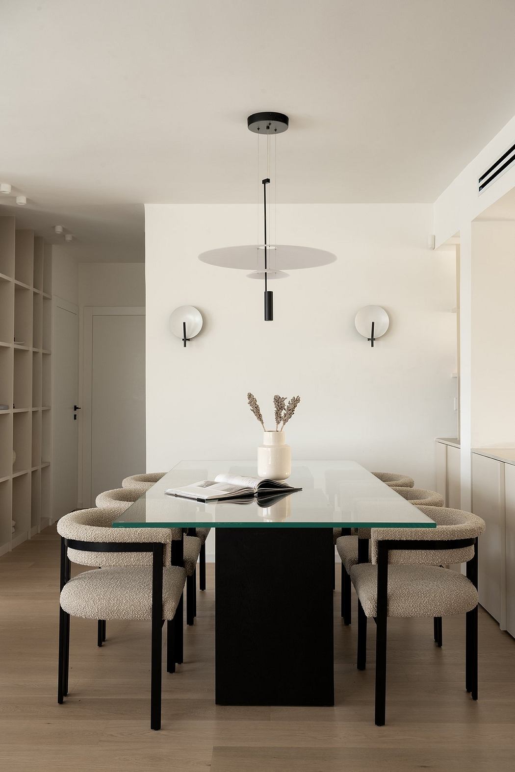 A modern dining area with a glass-topped table, beige upholstered chairs, and minimalist pendant lighting.
