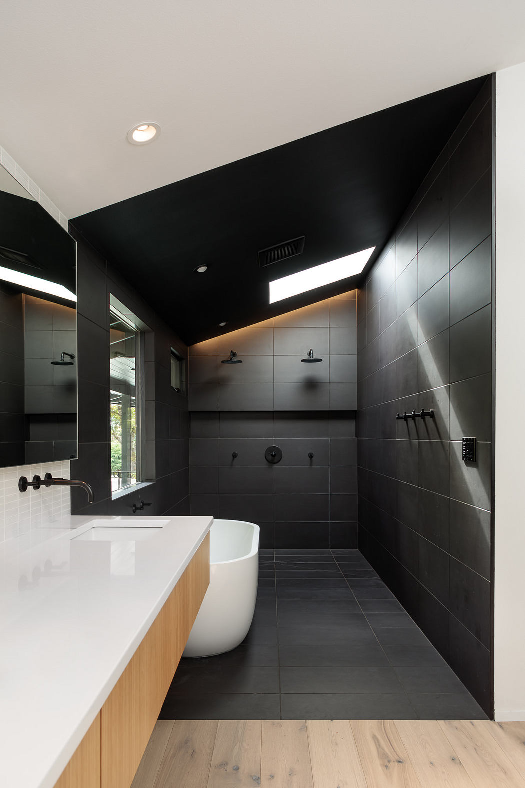 Modern bathroom with black tiled walls, freestanding bathtub, and wooden vanity.