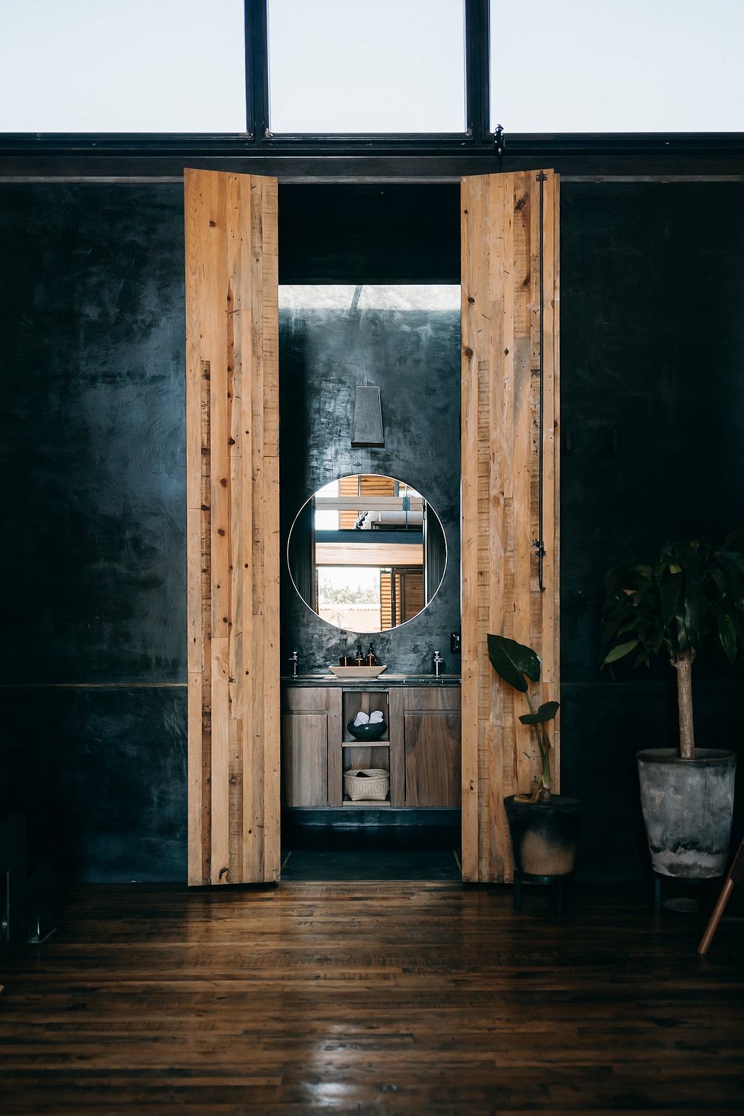 A dark, rustic bathroom with wooden panels, a round mirror, and potted plants.