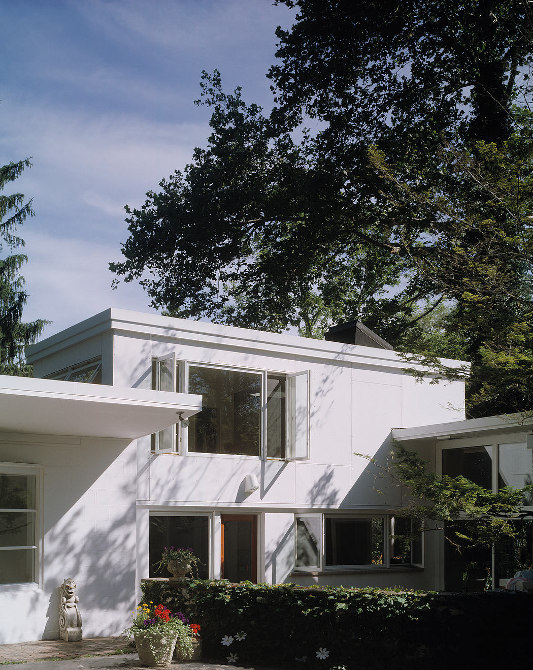 A modern, white, two-story home with geometric shapes, large windows, and a lush garden setting.