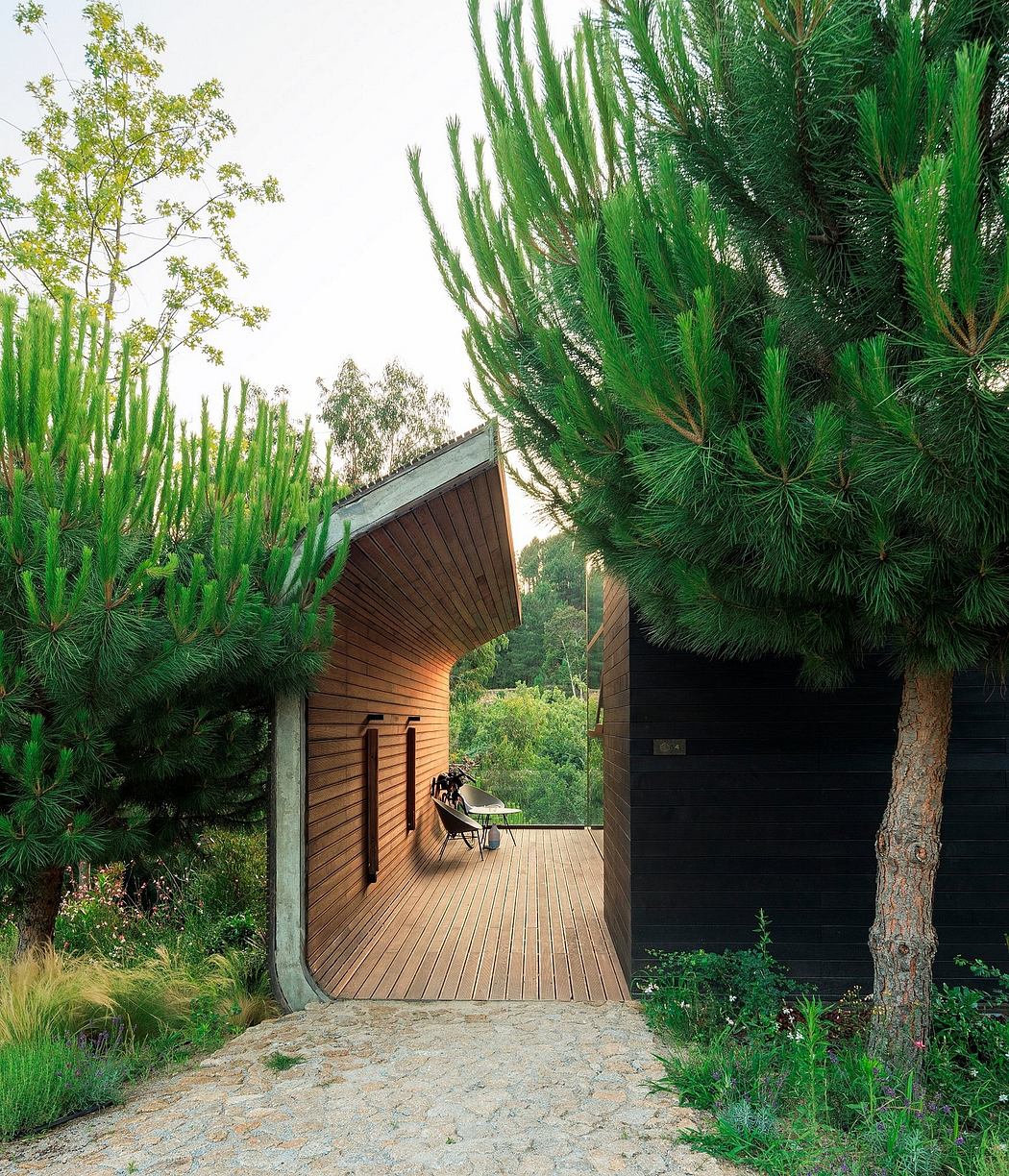 A wooden cabin with a curved roof sits amidst lush foliage, featuring a stone pathway leading to a wooden deck.
