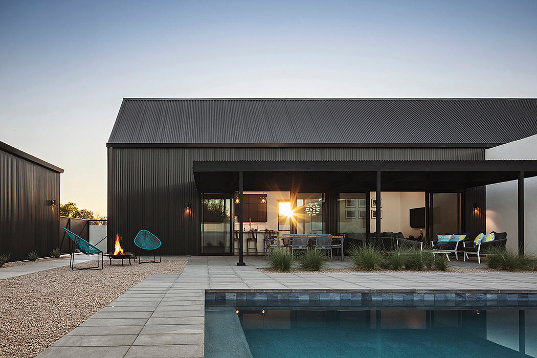 A modern, black metal-clad structure with a large covered patio and pool at sunset.