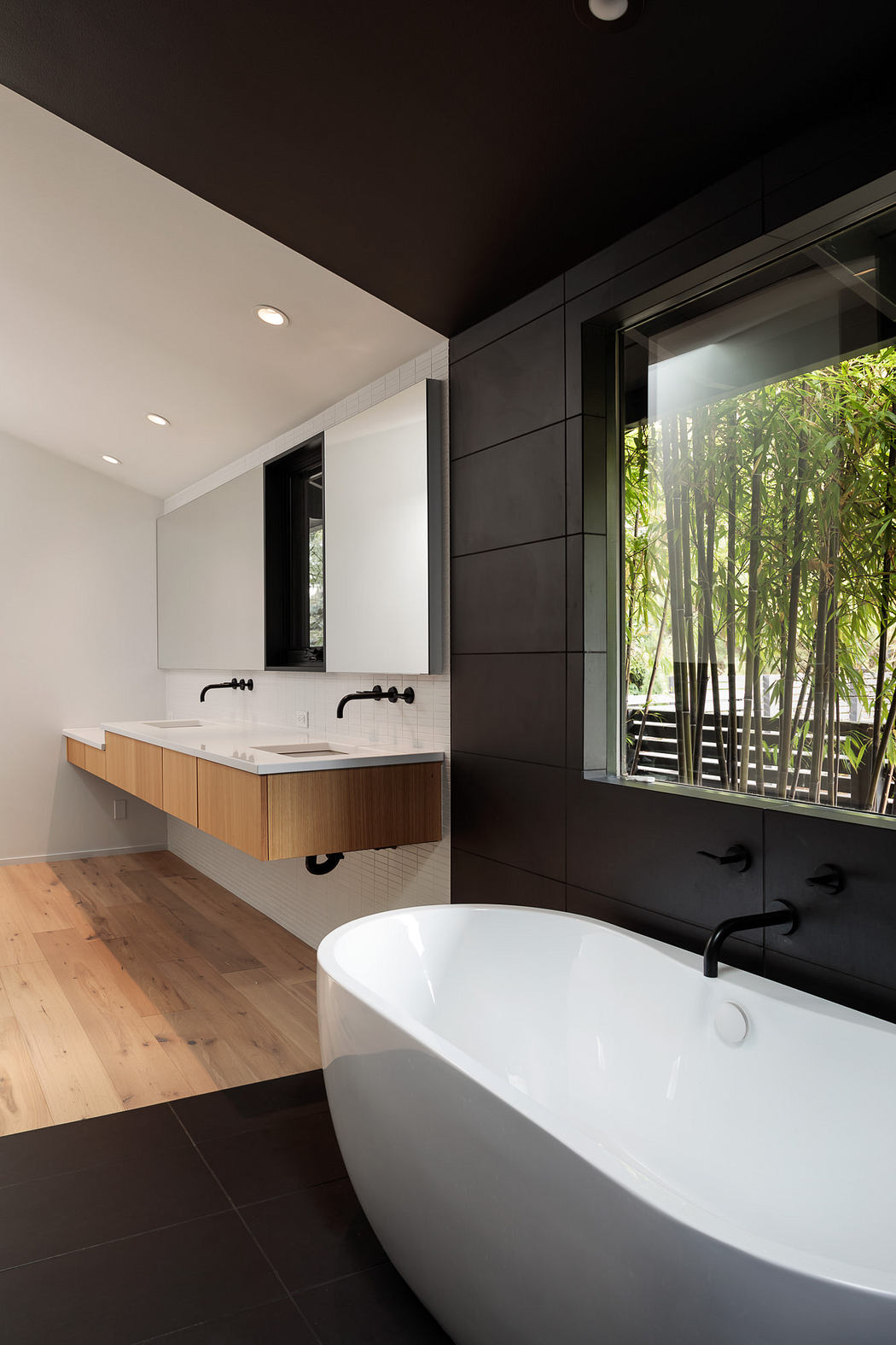 A modern, minimalist bathroom with a freestanding tub, wooden vanity, and large window overlooking greenery.
