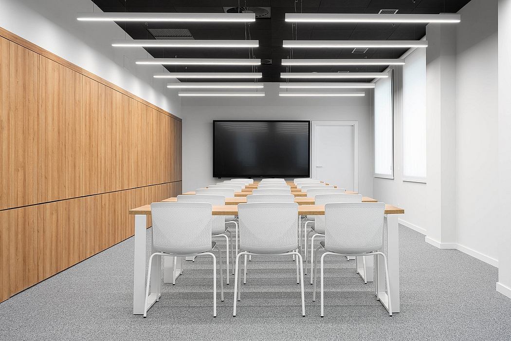Modern conference room with wooden walls, overhead lighting, and rows of white chairs facing a large display screen.