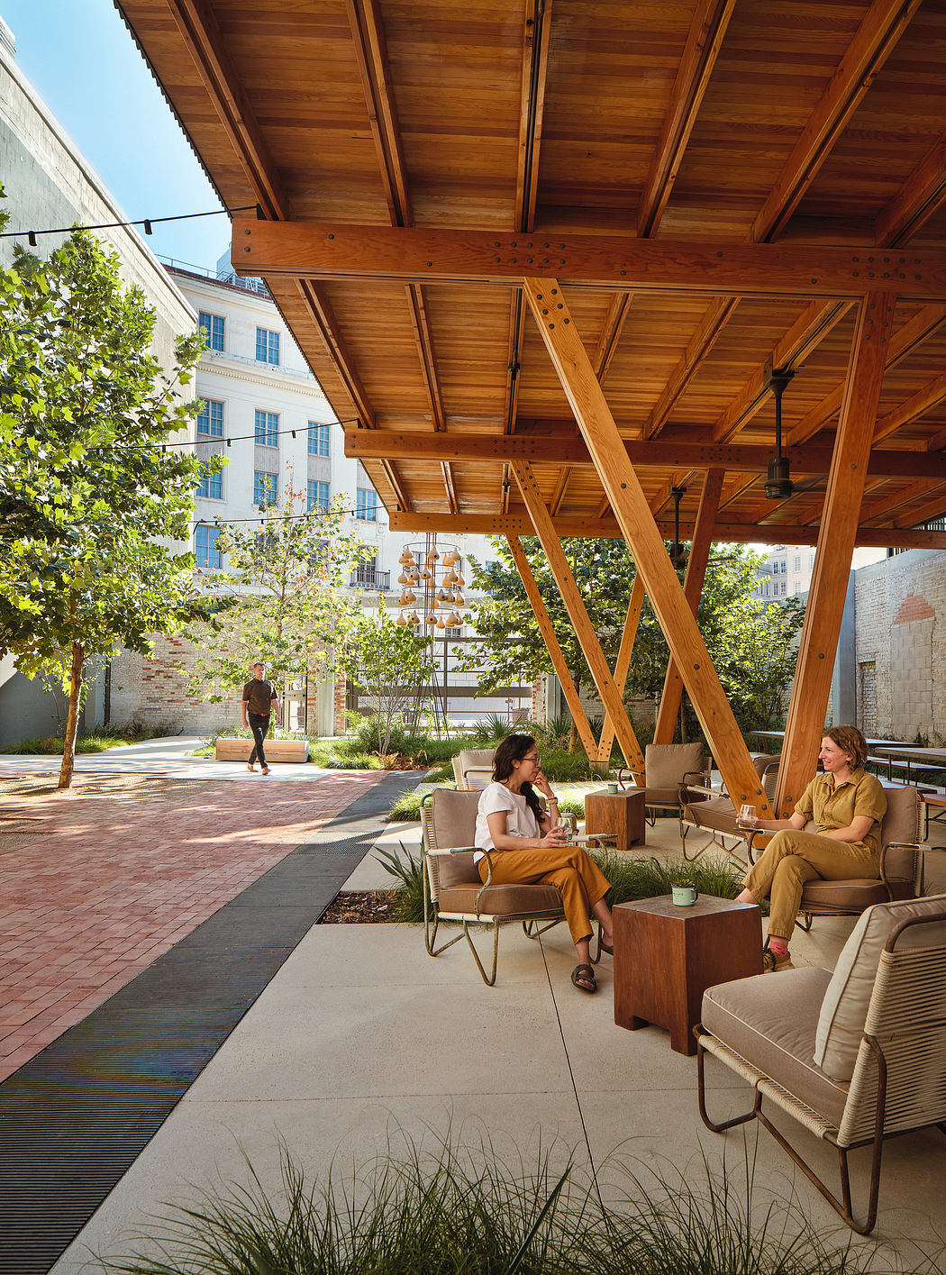 Covered wooden patio with seating area, surrounded by greenery and brick buildings.
