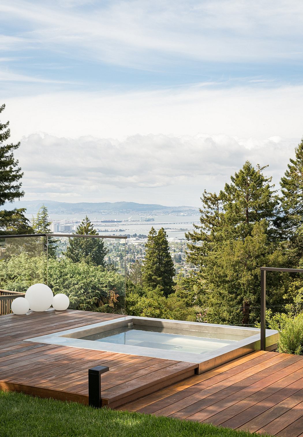 A modern wooden deck with a glass-enclosed hot tub, surrounded by lush greenery and a panoramic city view.
