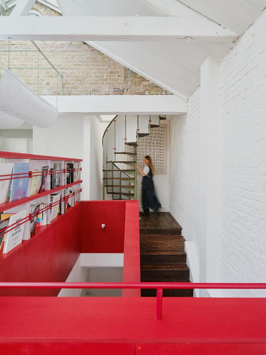 A modern, minimalist interior with exposed brick walls, red metal shelving, and a spiral staircase.