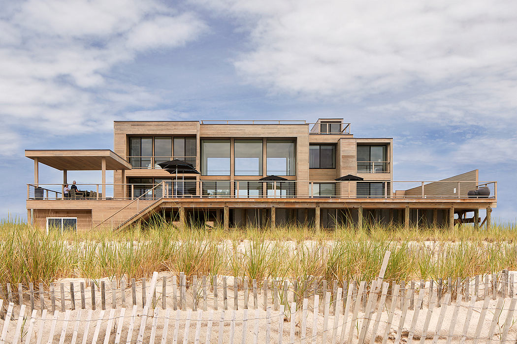 Contemporary beach house with wooden decks, balconies, and large windows overlooking the grass-covered dunes.