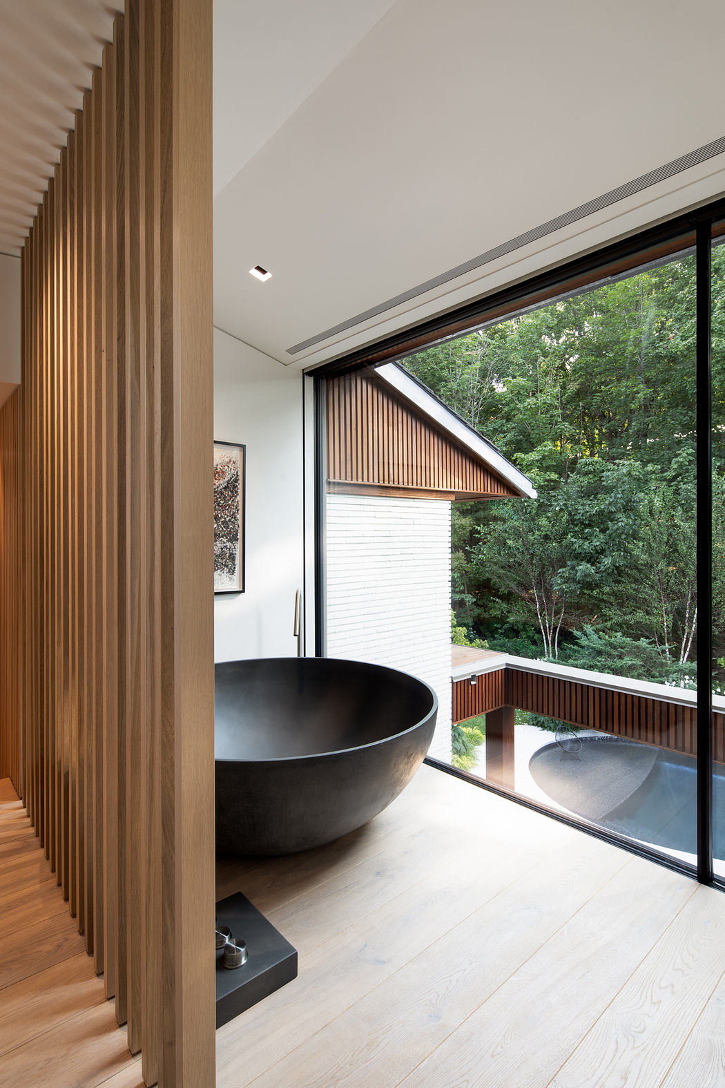A modern bathroom with a circular black tub, large windows overlooking nature, and wooden accents.