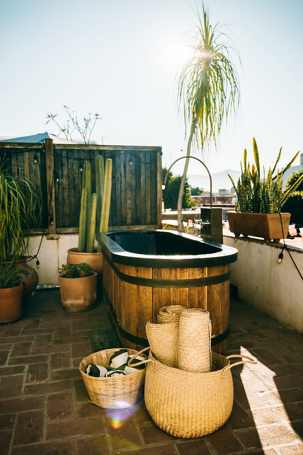 A cozy outdoor patio with a wooden hot tub, potted plants, and woven baskets, creating a soothing ambiance.