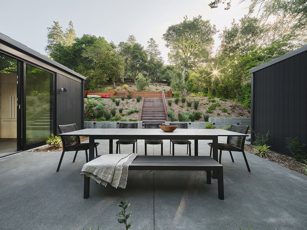 A modern outdoor dining area with a large table, bench, and chairs set against a lush, forested backdrop.